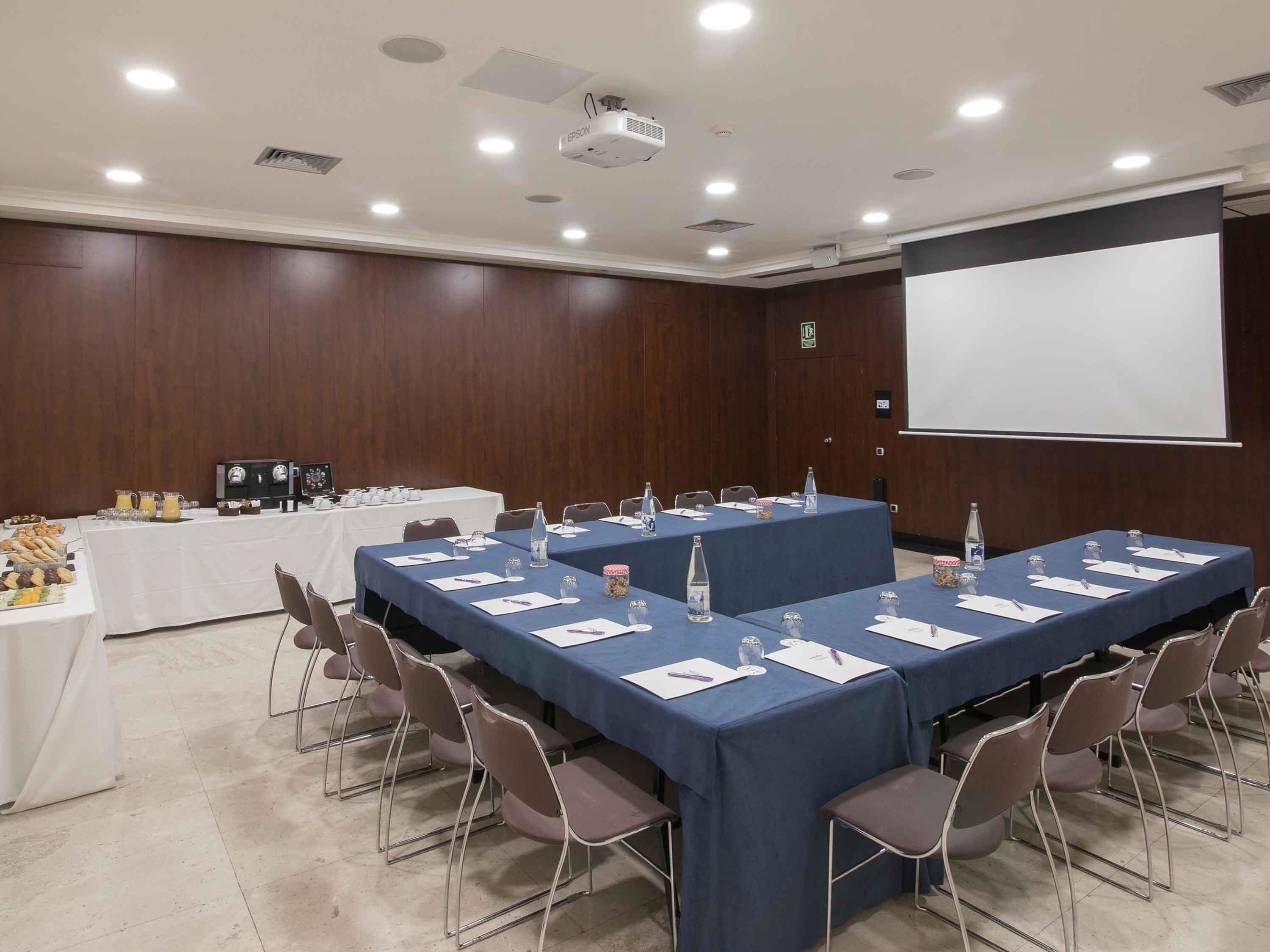 Una sala de conferencias con una larga mesa, sillas, un proyector y una pantalla.