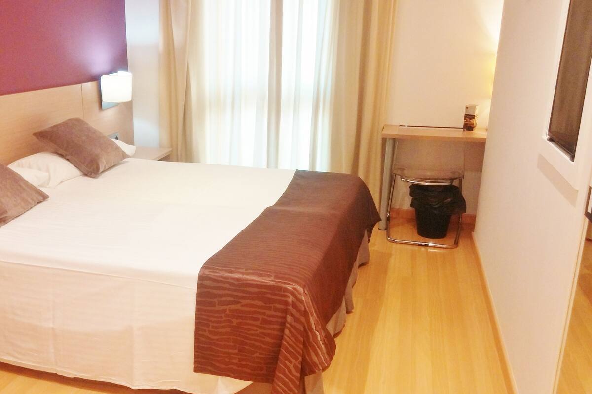 Quarto de hotel com uma cama grande, mesa de cabeceira, escrivaninha e uma cadeira.