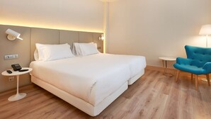 A hotel room with a large bed, bedside tables, a blue armchair, and a small table with a lamp. - NH Bilbao Deusto (Bilbao)