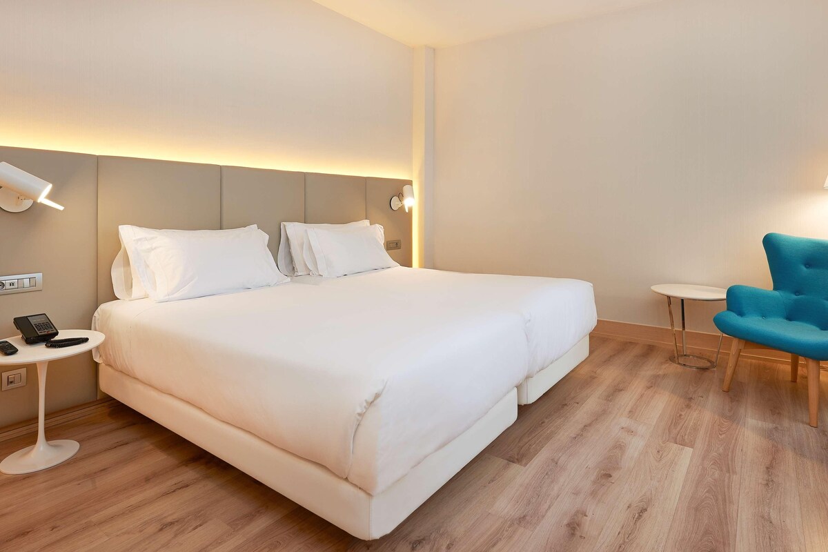 A hotel room with a large bed, bedside tables, a blue armchair, and a small table with a lamp.