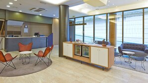 A modern reception area with a desk, chairs, a sofa, and a bar area with a refrigerator and glassware. - NH Bilbao Deusto (Bilbao)