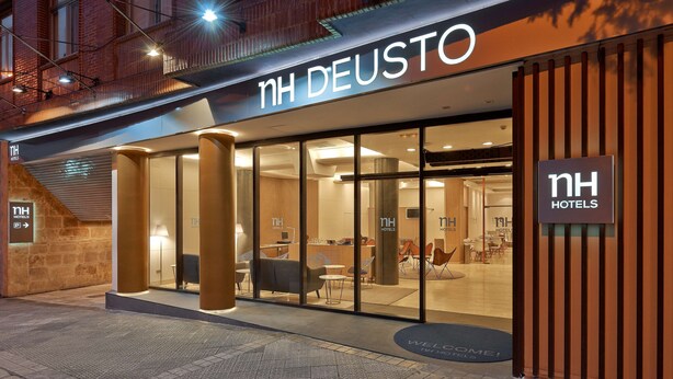 A hotel entrance with a modern design, featuring glass doors, a reception area, and a visible seating area inside. - NH Bilbao Deusto (Bilbao)