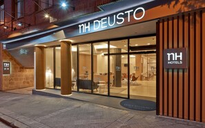 A hotel entrance with a modern design, featuring glass doors, a reception area, and a visible seating area inside. - NH Bilbao Deusto (Bilbao)