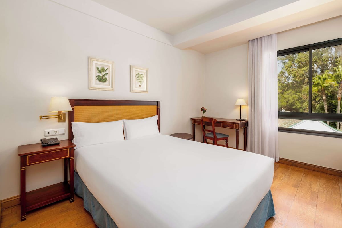 A hotel room with a large bed, a desk, two lamps, and a window with a view of trees.