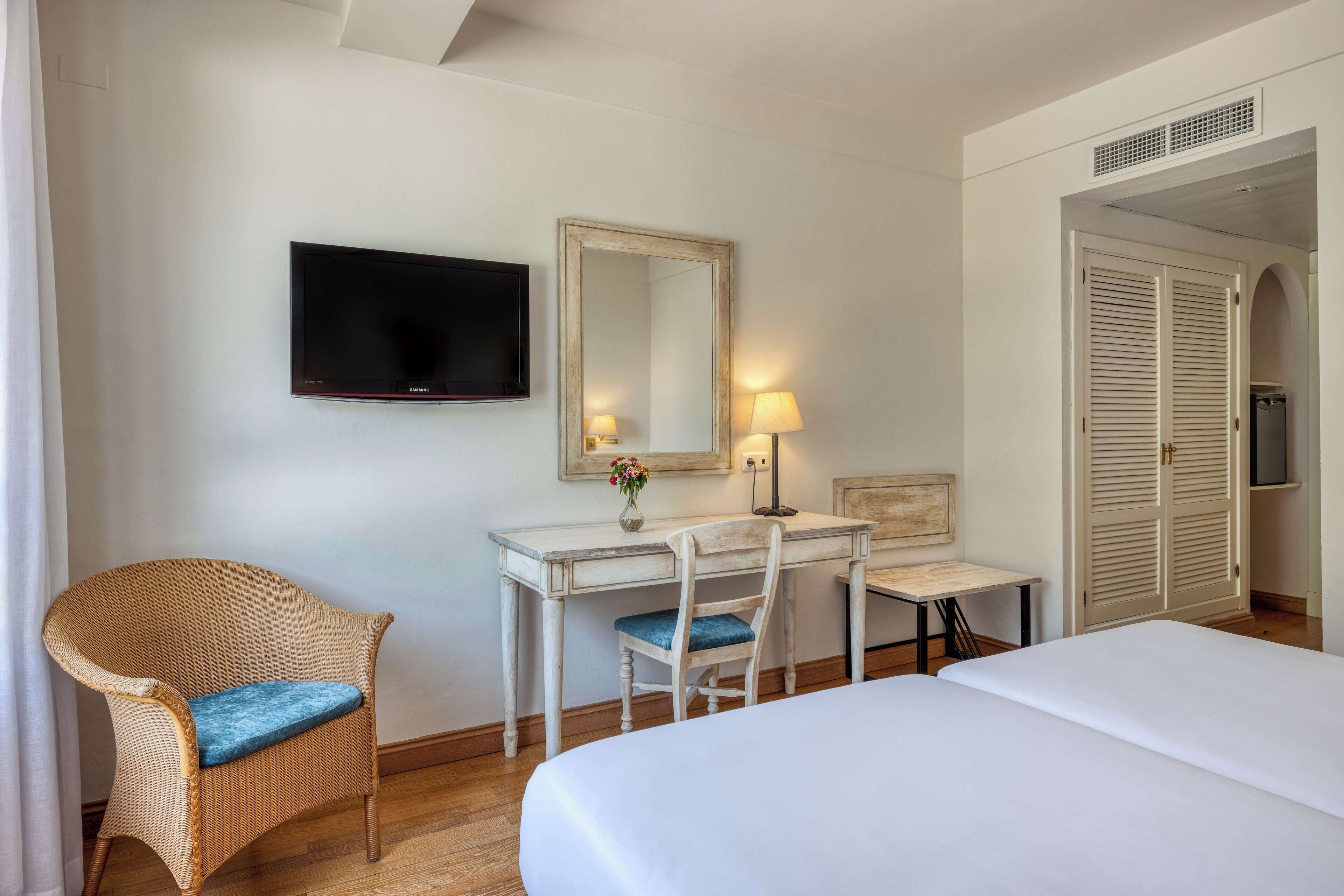 A hotel room with a bed, a desk with a chair, a television, a mirror, and a wardrobe.
