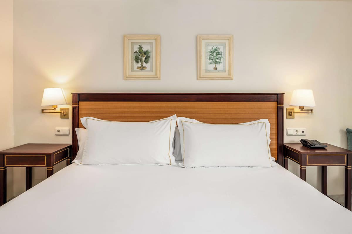 A neatly made bed with white linens and two pillows, flanked by two bedside tables with lamps and framed botanical prints on the wall above.