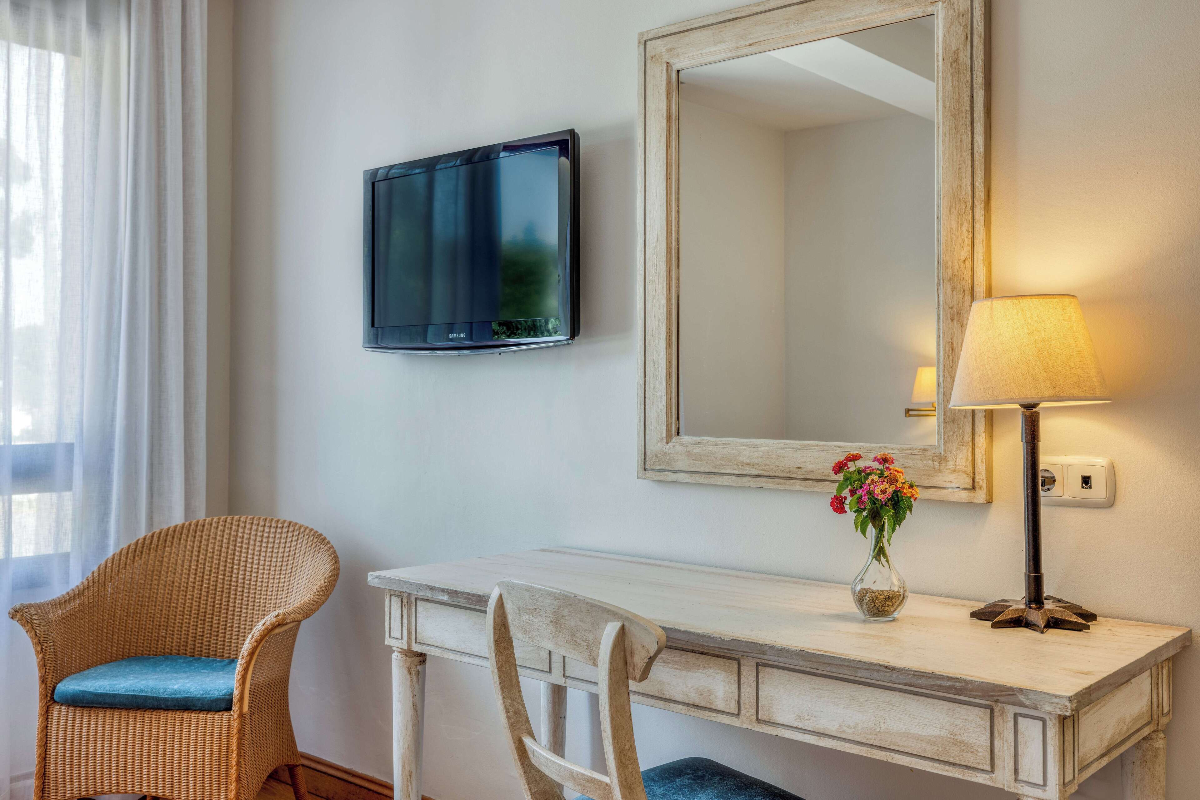 A room with a white desk, two chairs, a television, a mirror, and a lamp.
