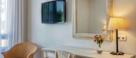 A room with a white desk, two chairs, a television, a mirror, and a lamp.