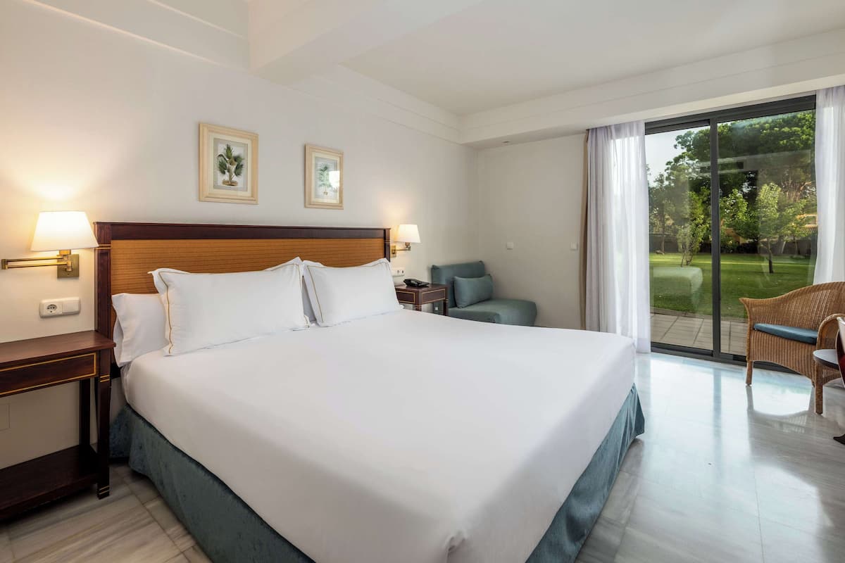 A hotel room with a large bed, two wall-mounted lamps, a nightstand, a chair, and a view of a garden through a sliding glass door.