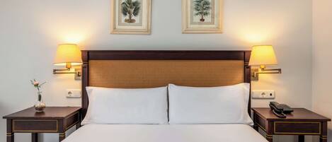 Habitación de hotel con una cama grande, dos mesitas de noche con lámparas y dos cuadros con motivos botánicos en la pared.