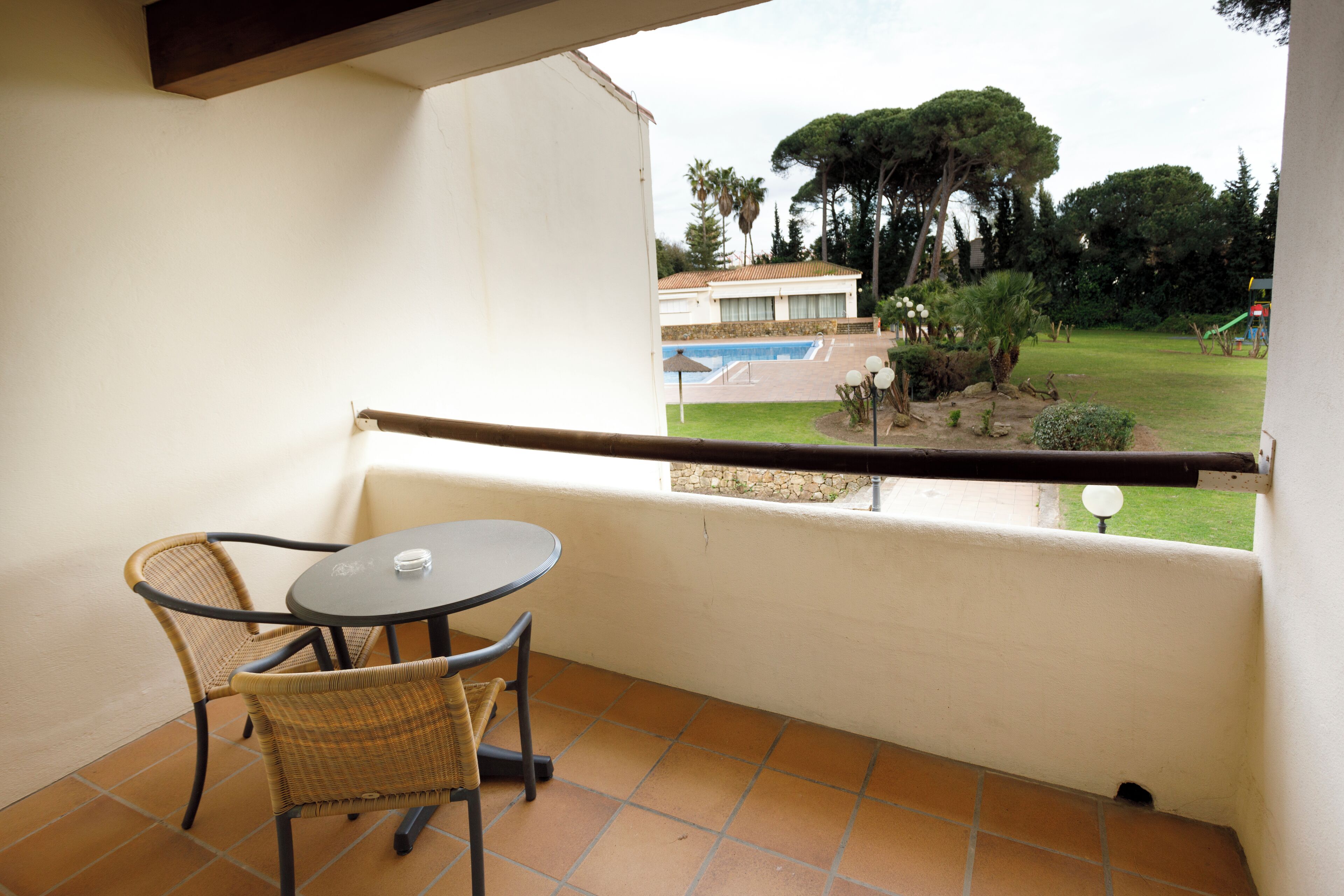 A balcony with a table and chairs, overlooking a swimming pool and a garden.