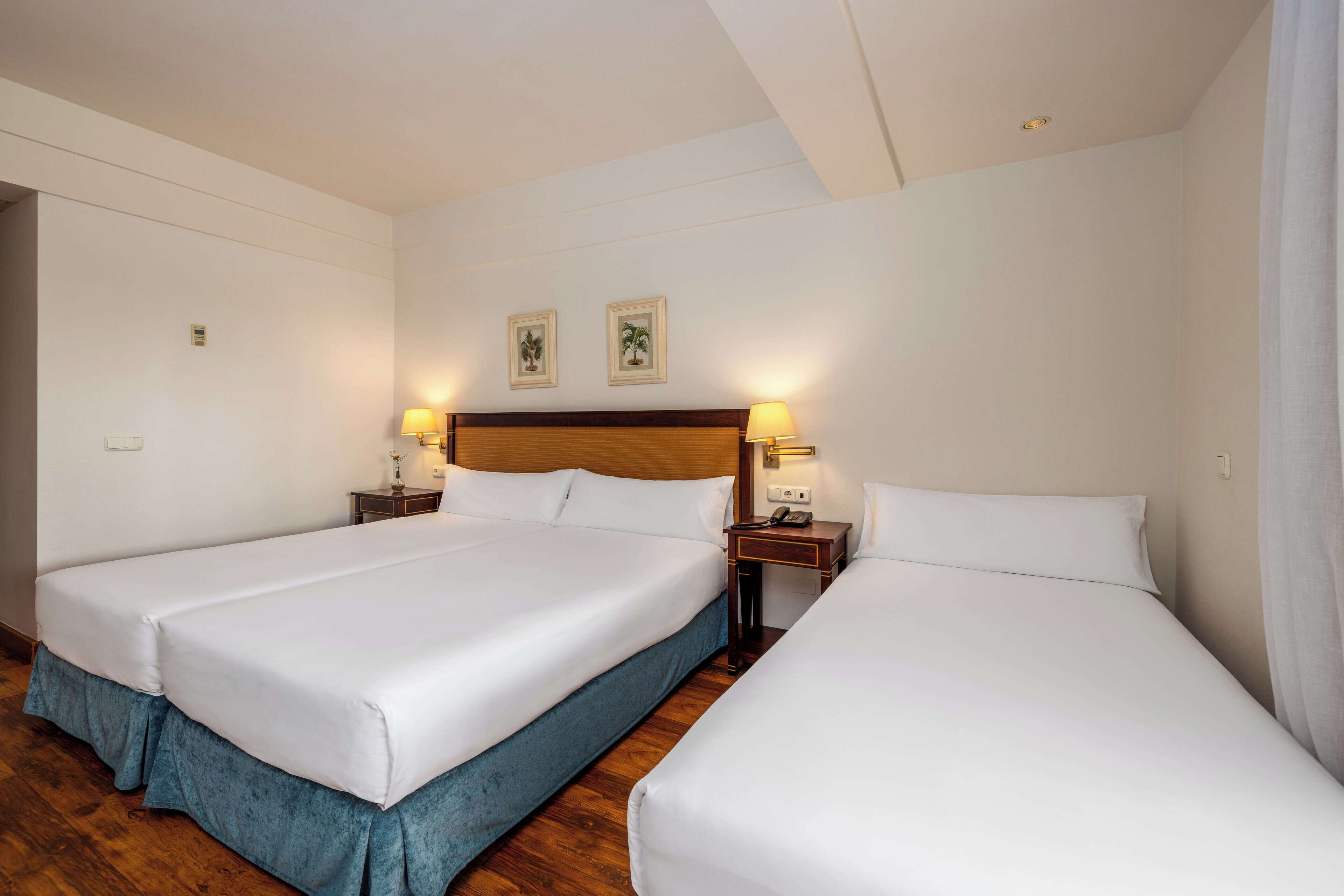 Una habitación de hotel con dos camas individuales, cabeceros de madera, mesitas de noche con lámparas e iluminación empotrada en el techo.