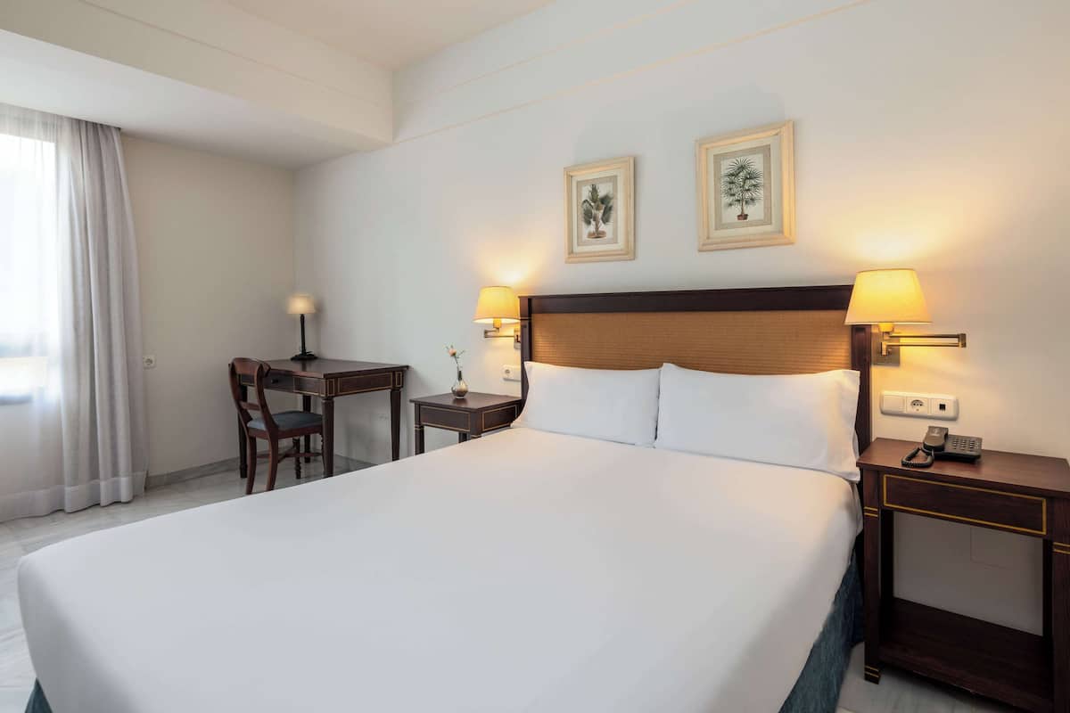 A hotel room with a large bed, two bedside lamps, a desk with a chair, and two framed pictures on the wall.