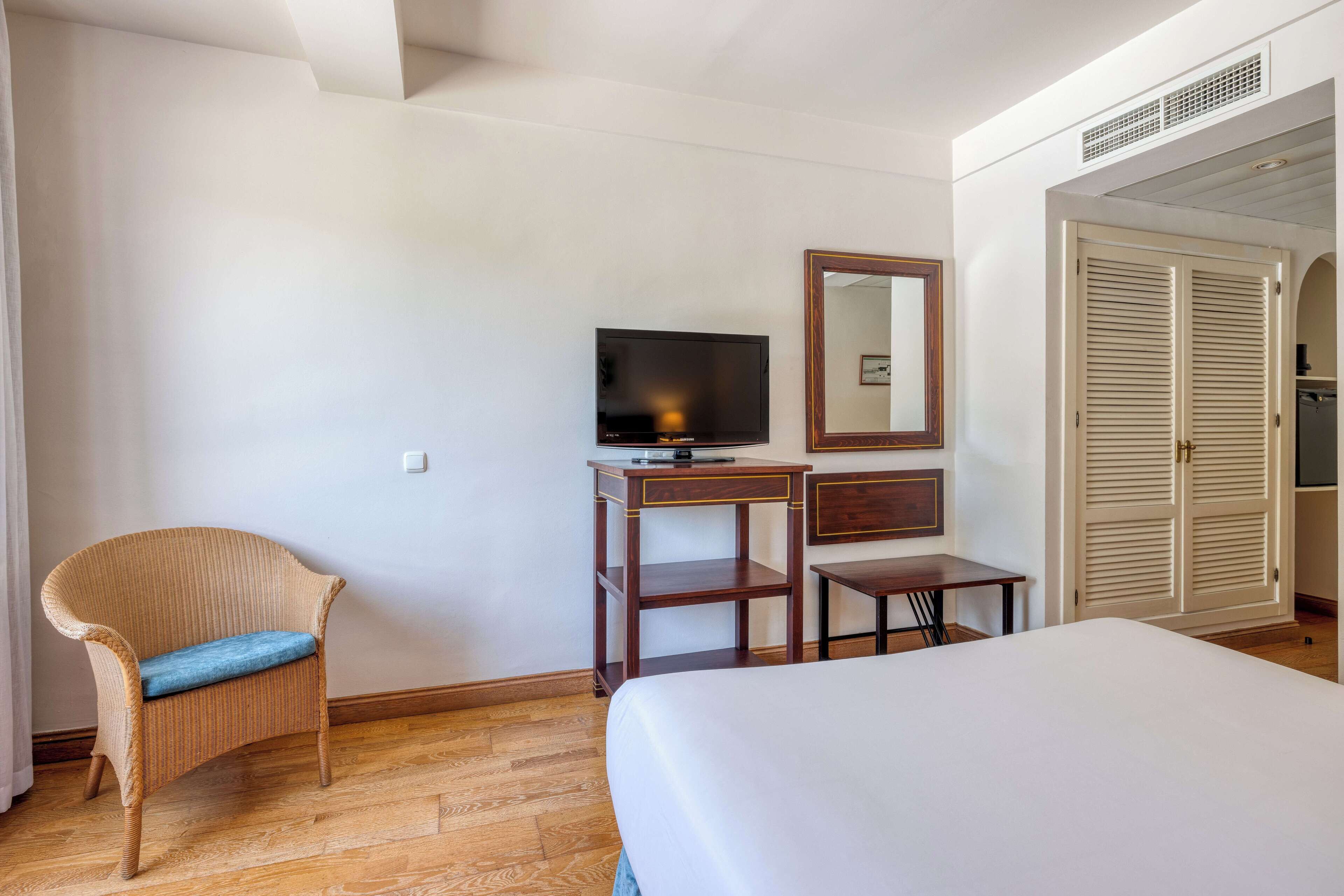 Ein Hotelzimmer mit einem Bett, einem Fernseher auf einem Holztisch, einem Korbstuhl mit Kissen, einem kleinen Tisch und einem Kleiderschrank.