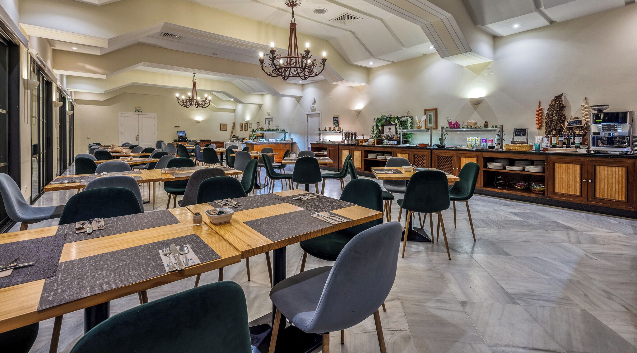 Un amplio comedor con múltiples mesas y sillas, una zona de bar central y una lámpara de araña moderna.