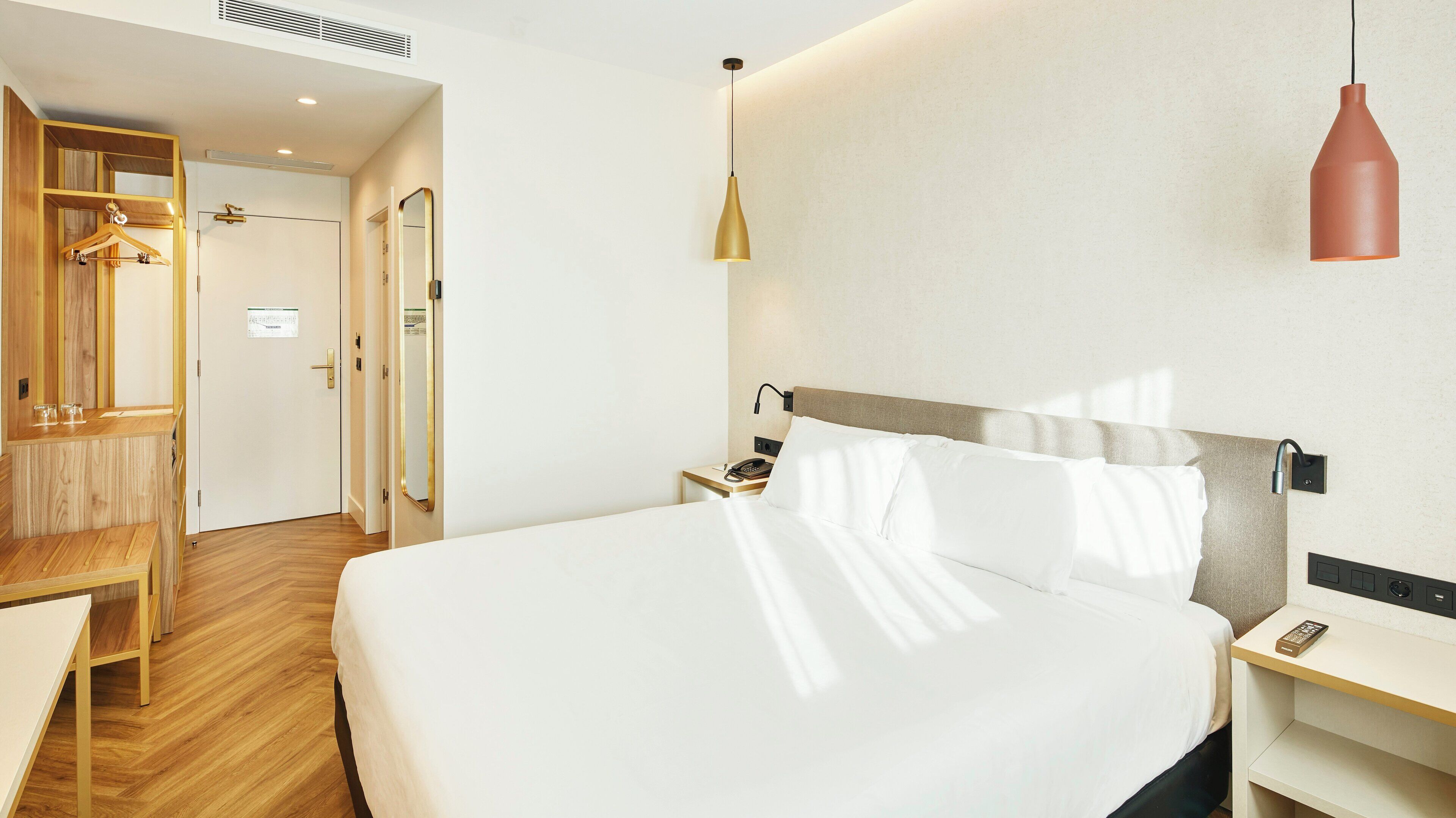 A hotel room with a large bed, bedside table, and a desk with a chair. There is a wardrobe with a mirror and a coat rack.