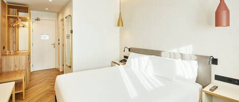 A hotel room with a large bed, bedside table, and a desk with a chair. There is a wardrobe with a mirror and a coat rack.