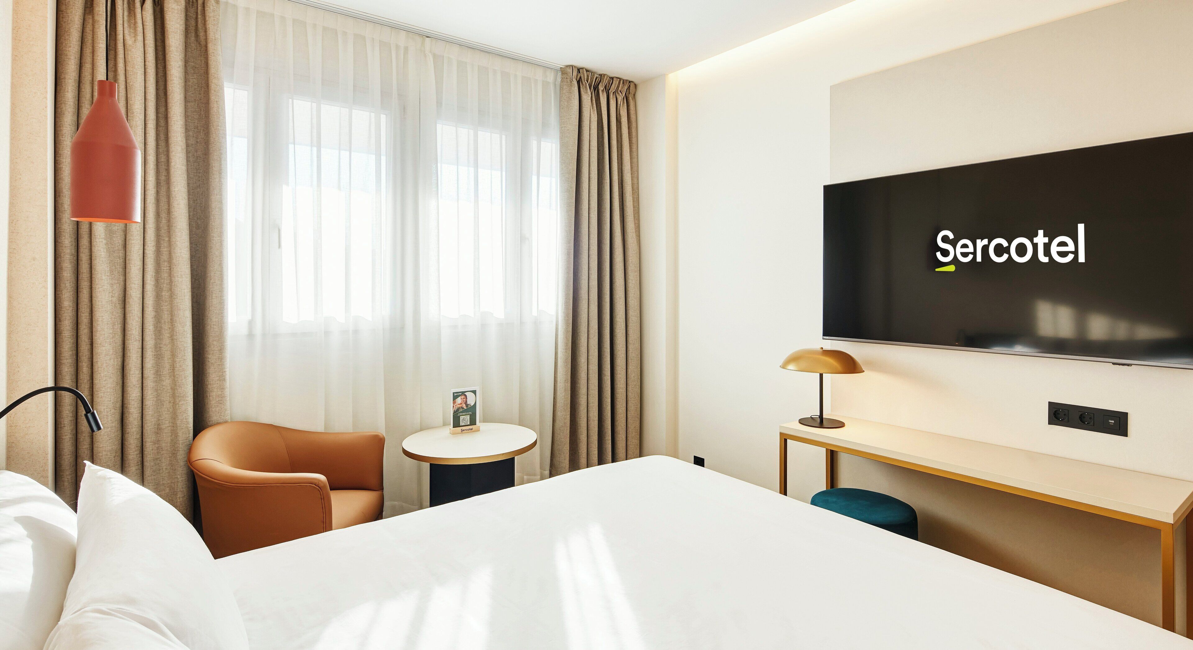A hotel room with a bed, a desk, a chair, a TV, and a window with curtains.