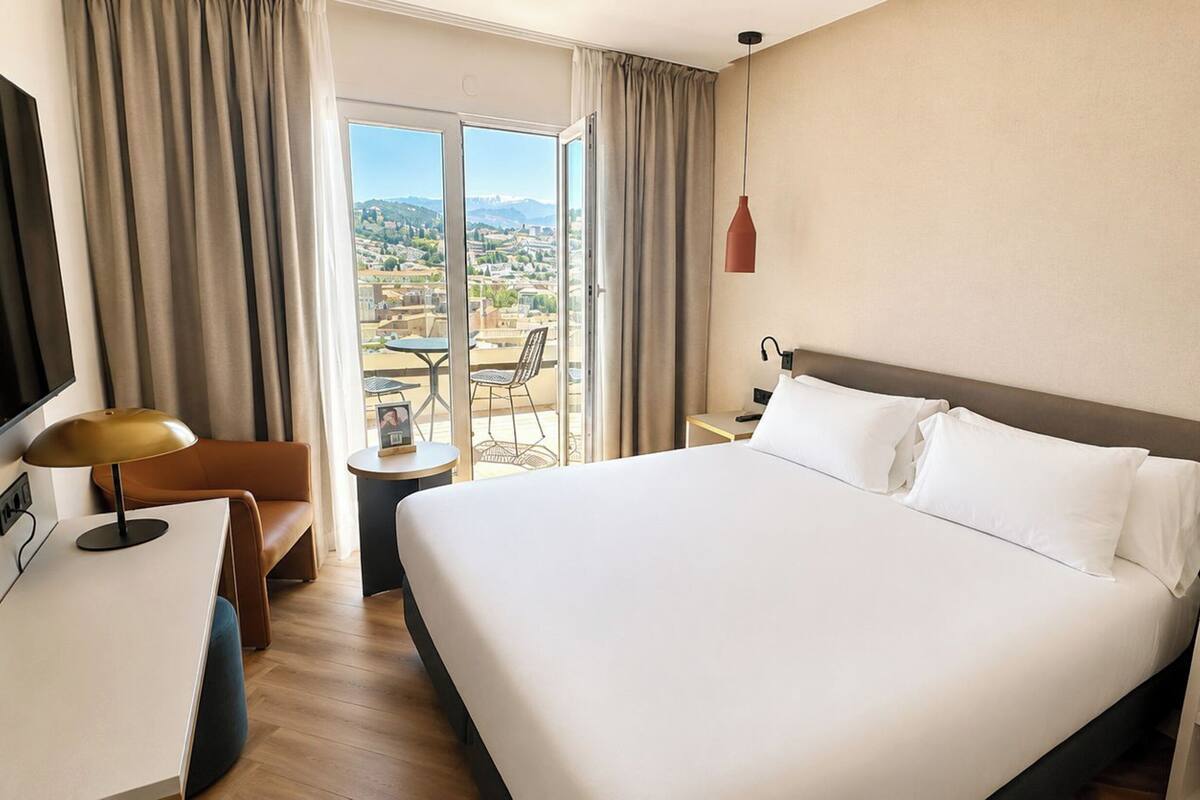A hotel room with a large bed, a desk, a chair, a TV, and a view of the city.