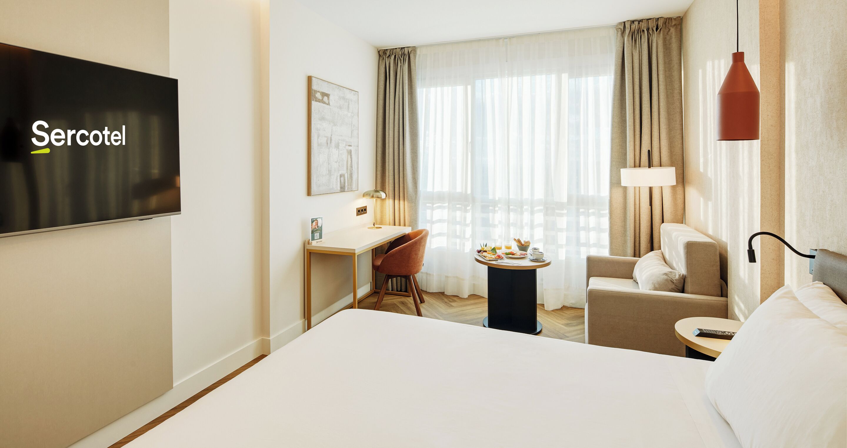 Hotelkamer met een bed, een bureau, een stoel, een klein tafeltje met een lamp en een televisietoestel waarop 'Sercotel' staat.