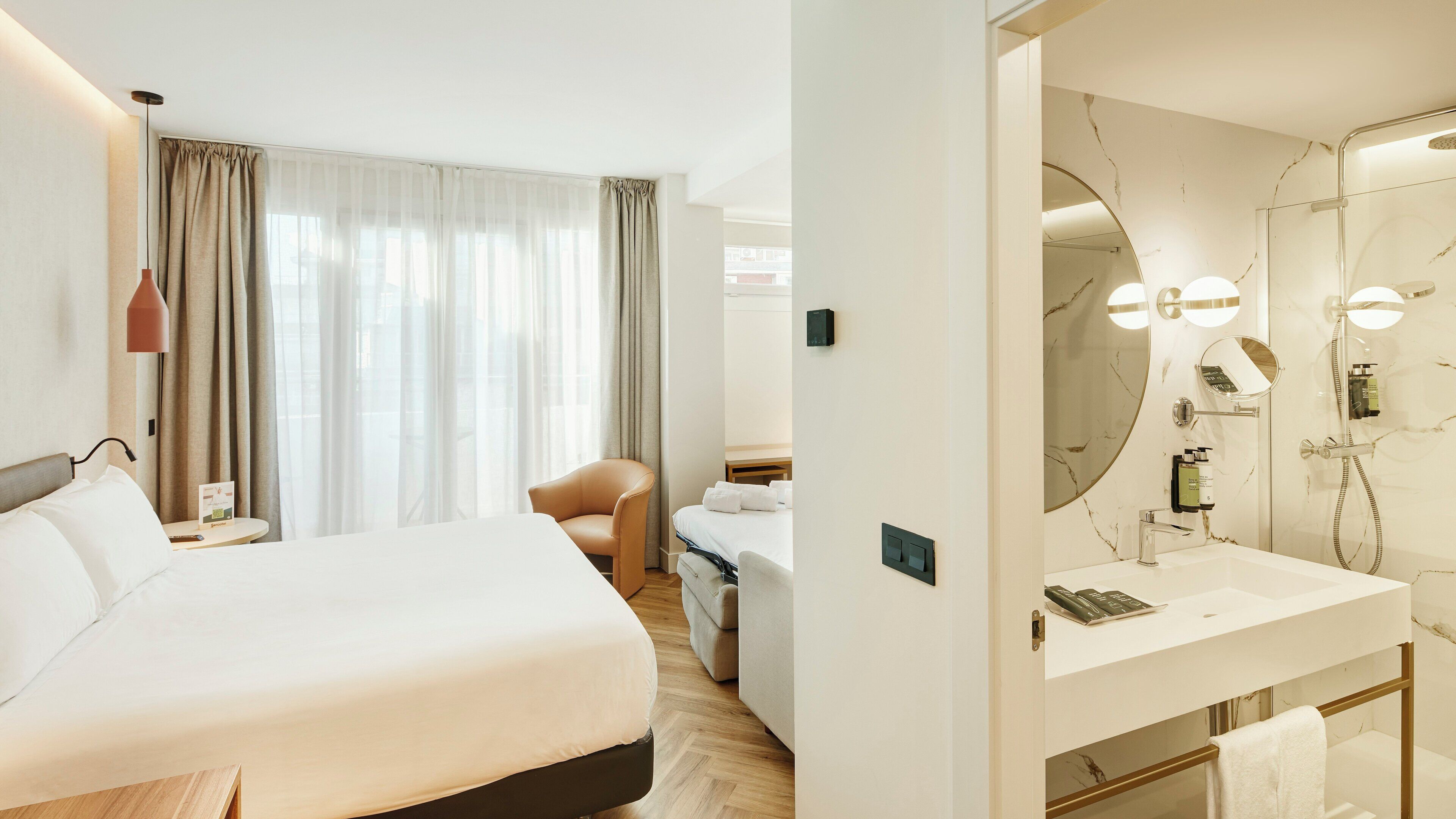 A hotel room with a bed, a chair, a desk with a lamp, a bathroom with a mirror, and a shower.
