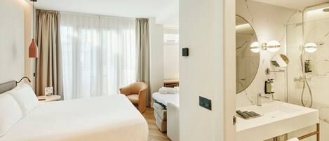 Habitación de hotel con cama, una silla, escritorio con lámpara, baño con espejo y ducha.