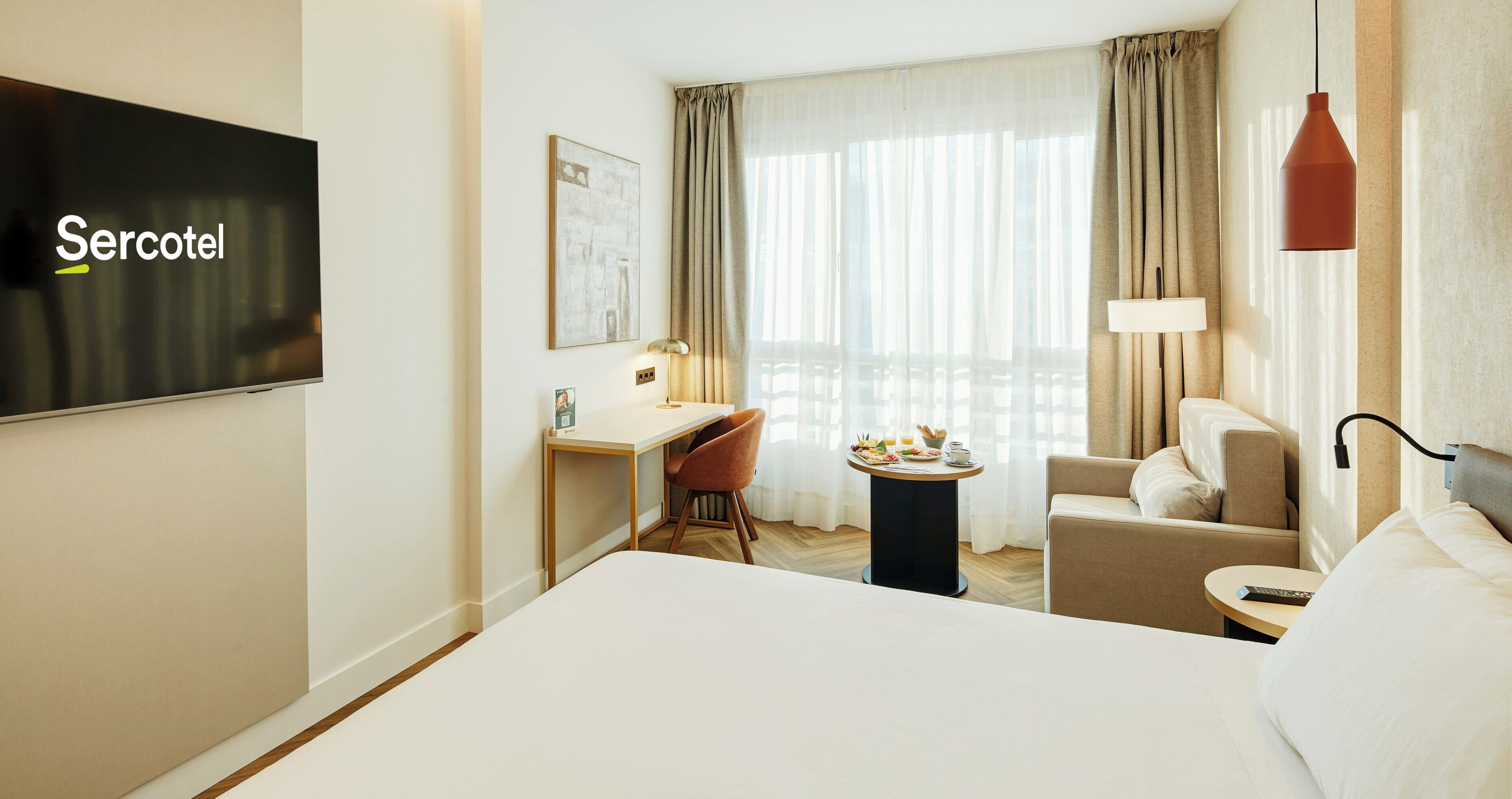 A hotel room with a bed, a desk, a chair, a small table with a lamp, and a television on the wall.