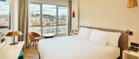 A hotel room with a large bed, a desk, a chair, a small table, and a view of a cityscape through large windows.