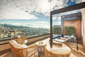 Un balcon situé dans un immeuble de grande hauteur, avec des meubles en osier, une table ronde sur laquelle sont disposées des boissons, et une vue sur la ville.