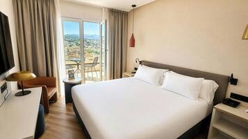 A hotel room with a large bed, a desk, a chair, a TV, a bedside table, a lamp, and a view of the city.