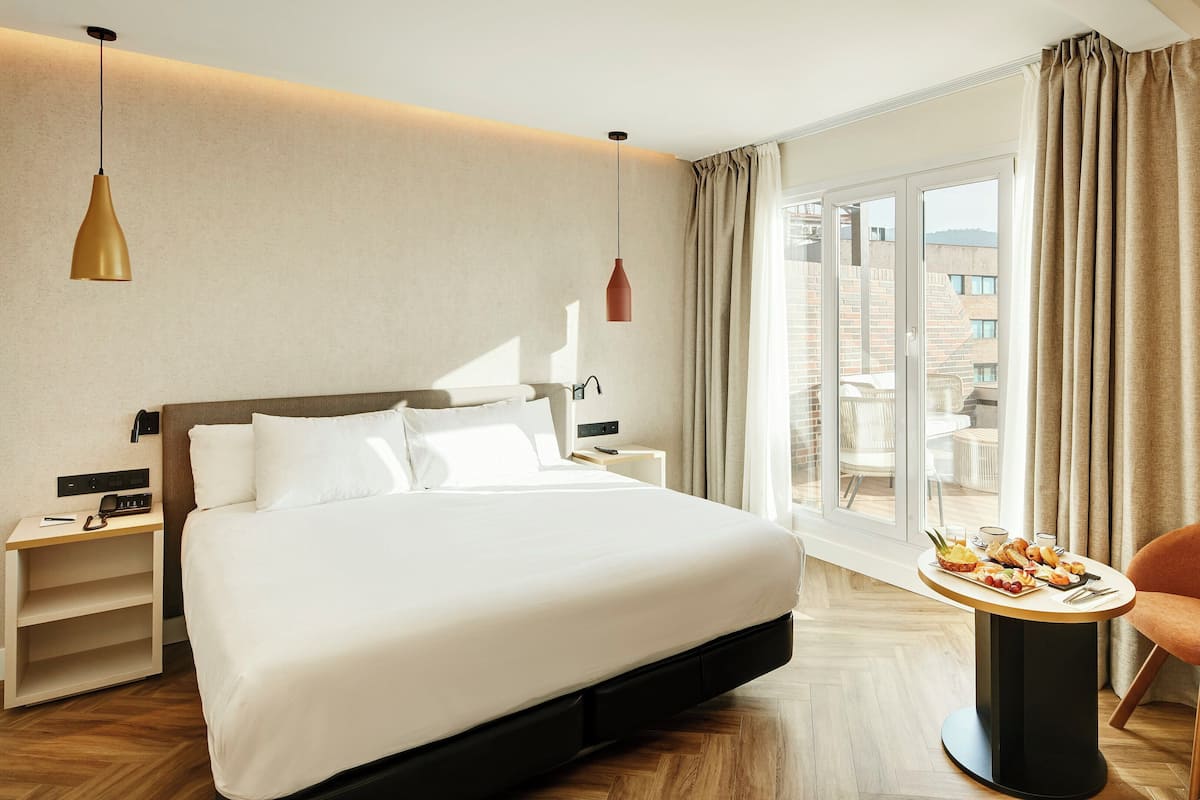 A hotel room with a large bed, a bedside table with a phone, a small table with breakfast, and a view of the city through the window.