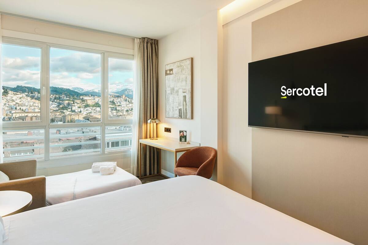 A hotel room with two beds, a desk, a chair, a TV, and a view of a cityscape.