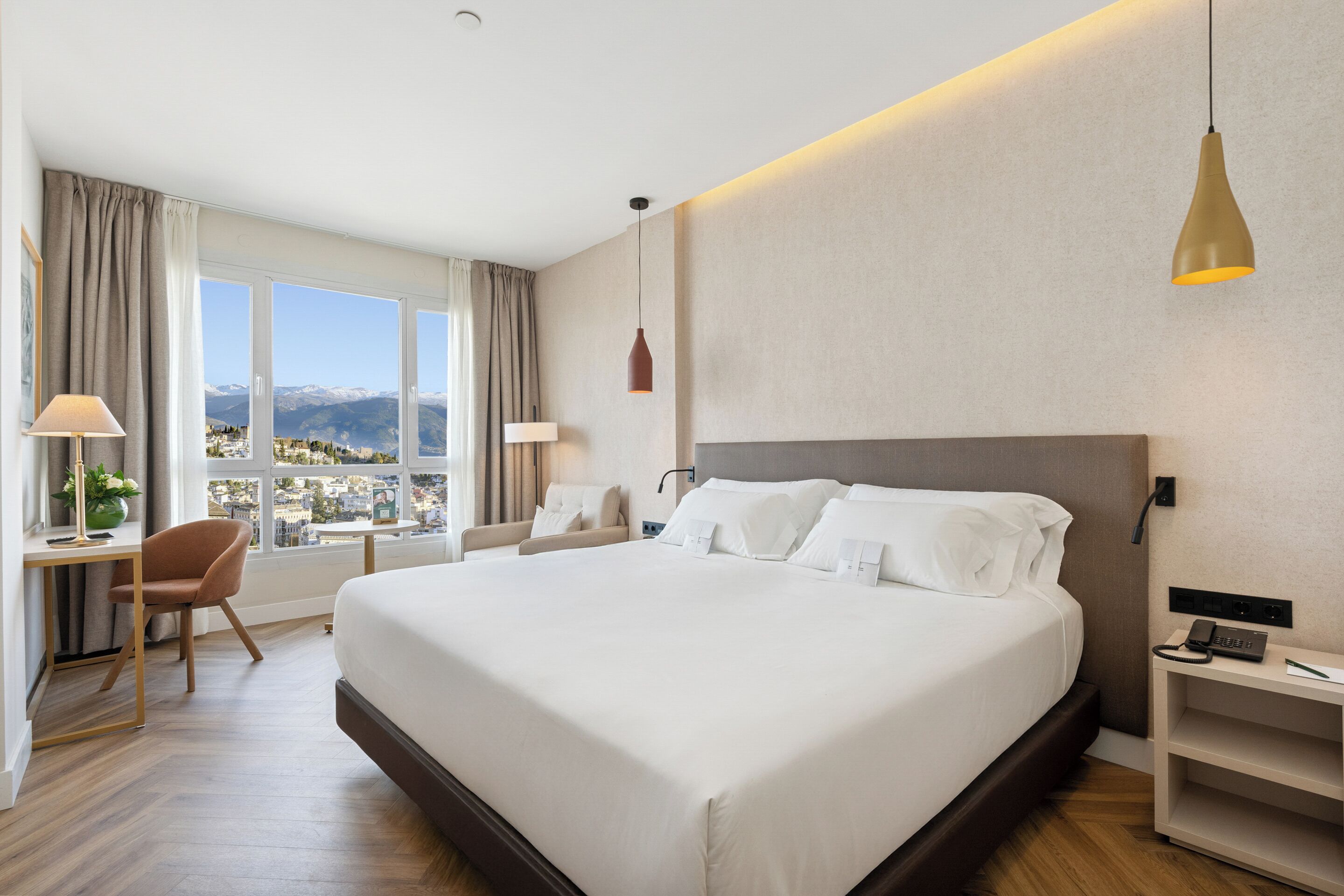 Habitación de hotel con una cama grande, un escritorio con lámpara, una silla, una mesita de noche con teléfono y vistas a las montañas por la ventana.
