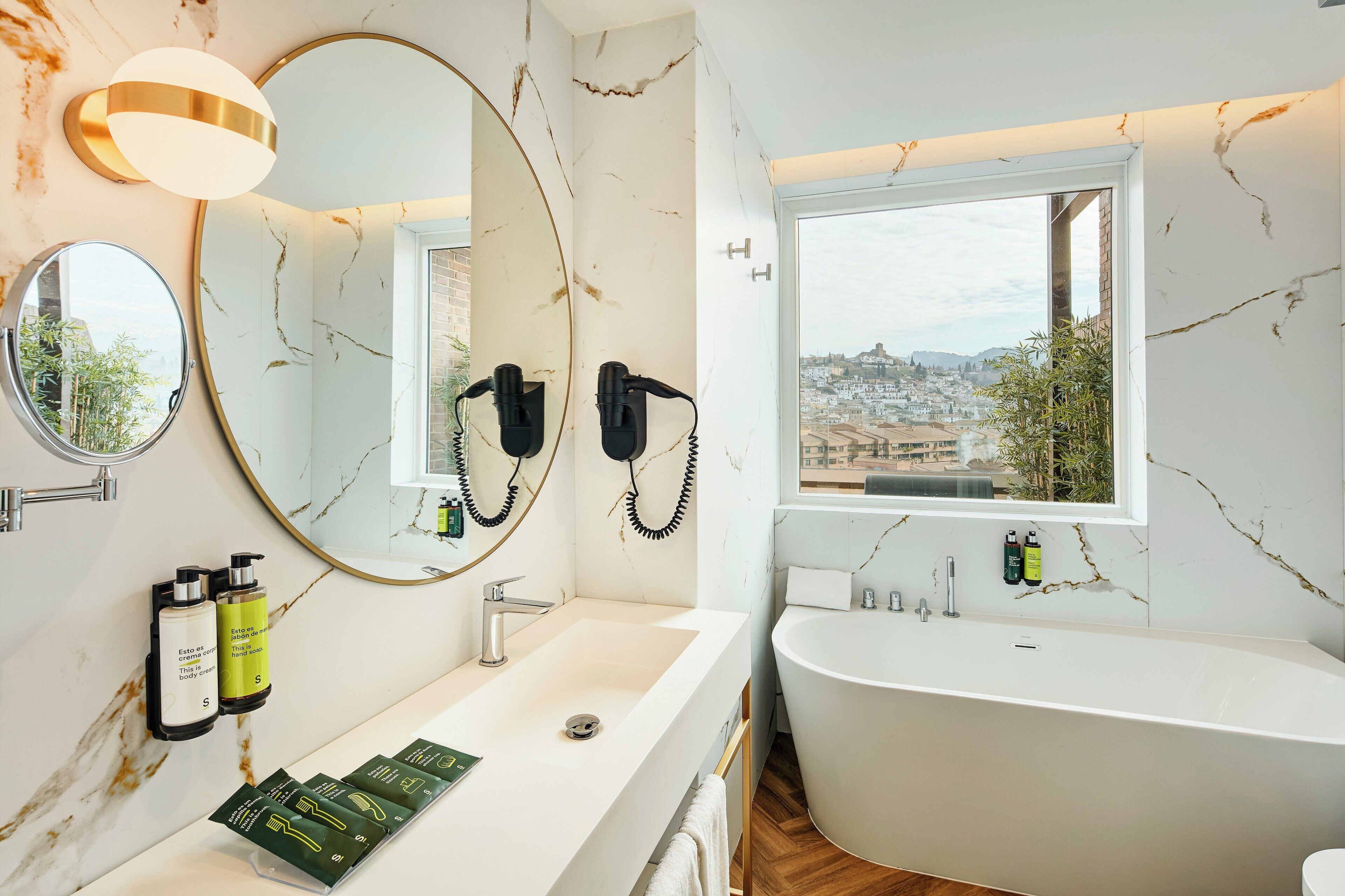 A modern bathroom with a large mirror, a round mirror, a sink, a bathtub, and a view of the city.