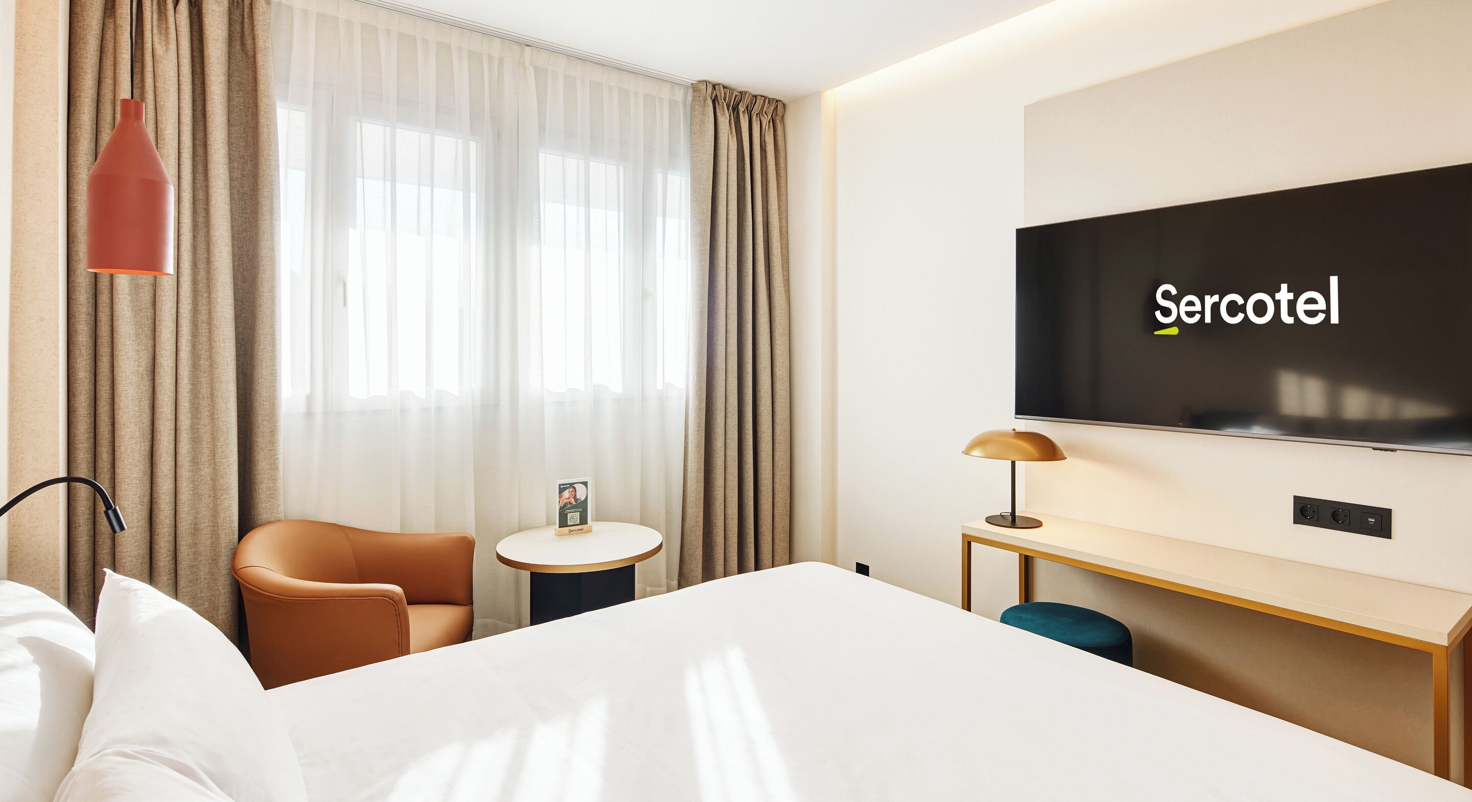 A hotel room with a bed, a desk, a chair, a TV, and a window with curtains.