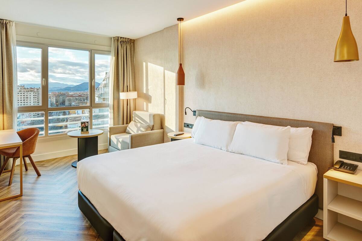 A hotel room with a large bed, a desk, a chair, a lamp, and a view of the city through the window.