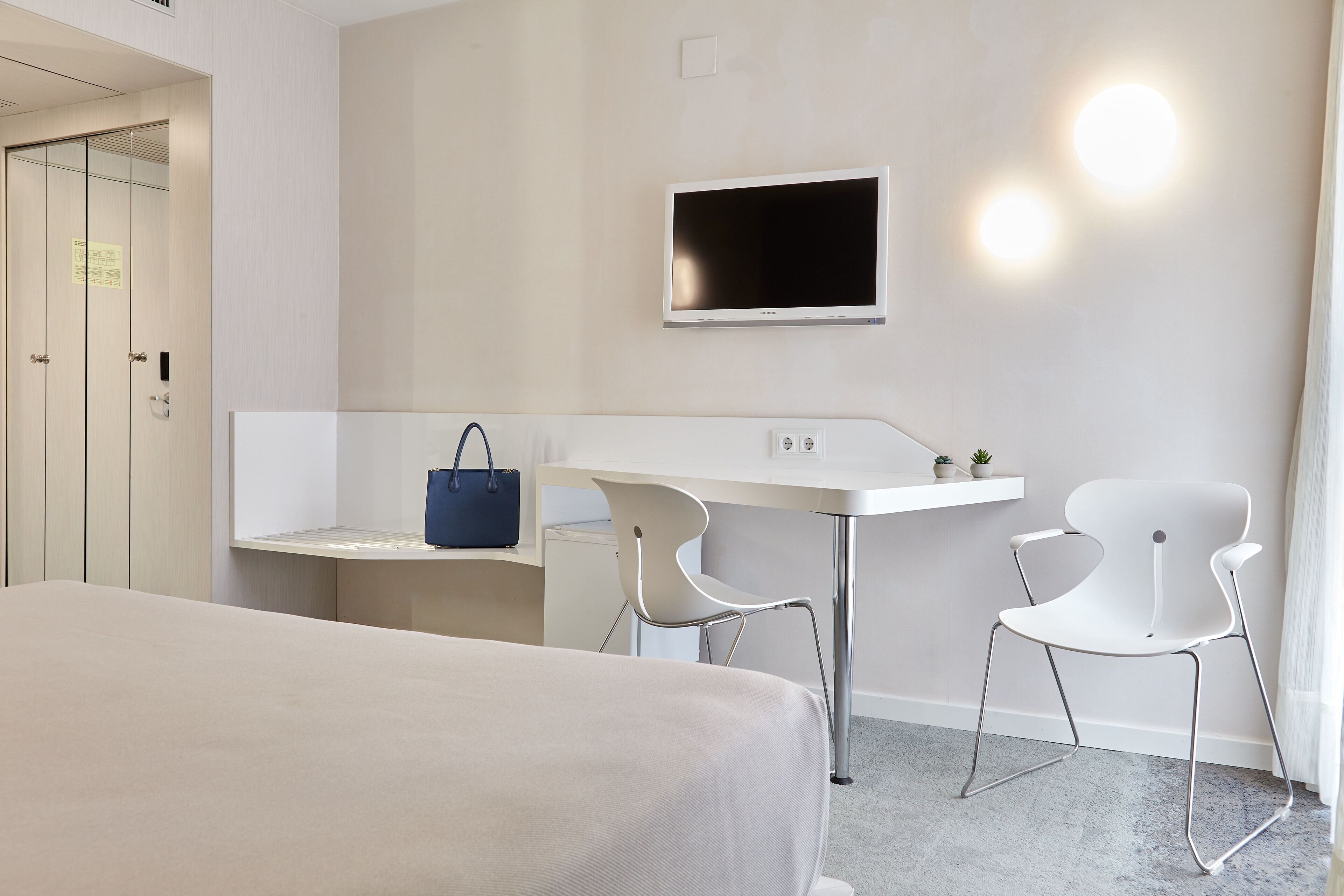 A hotel room with a bed, a desk, two chairs, a TV, and a blue bag on the desk.