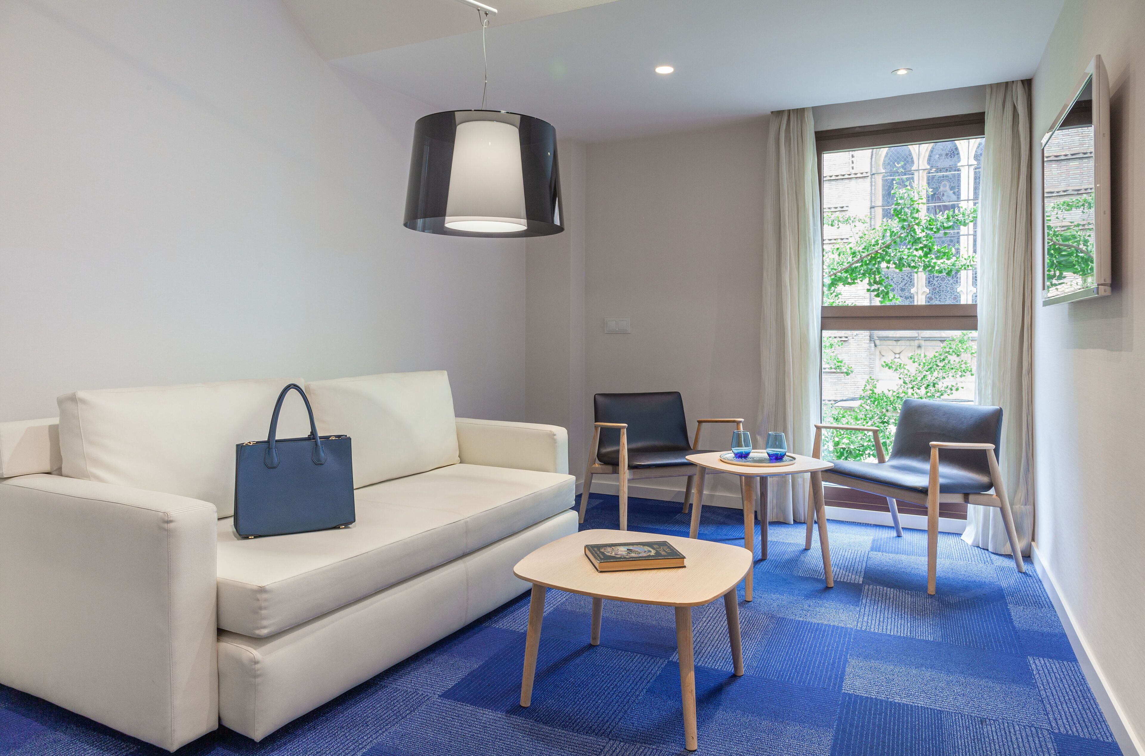 Een hotelkamer met een witte bank, een blauwe handtas, een klein houten tafeltje, een laptop en een stoel.