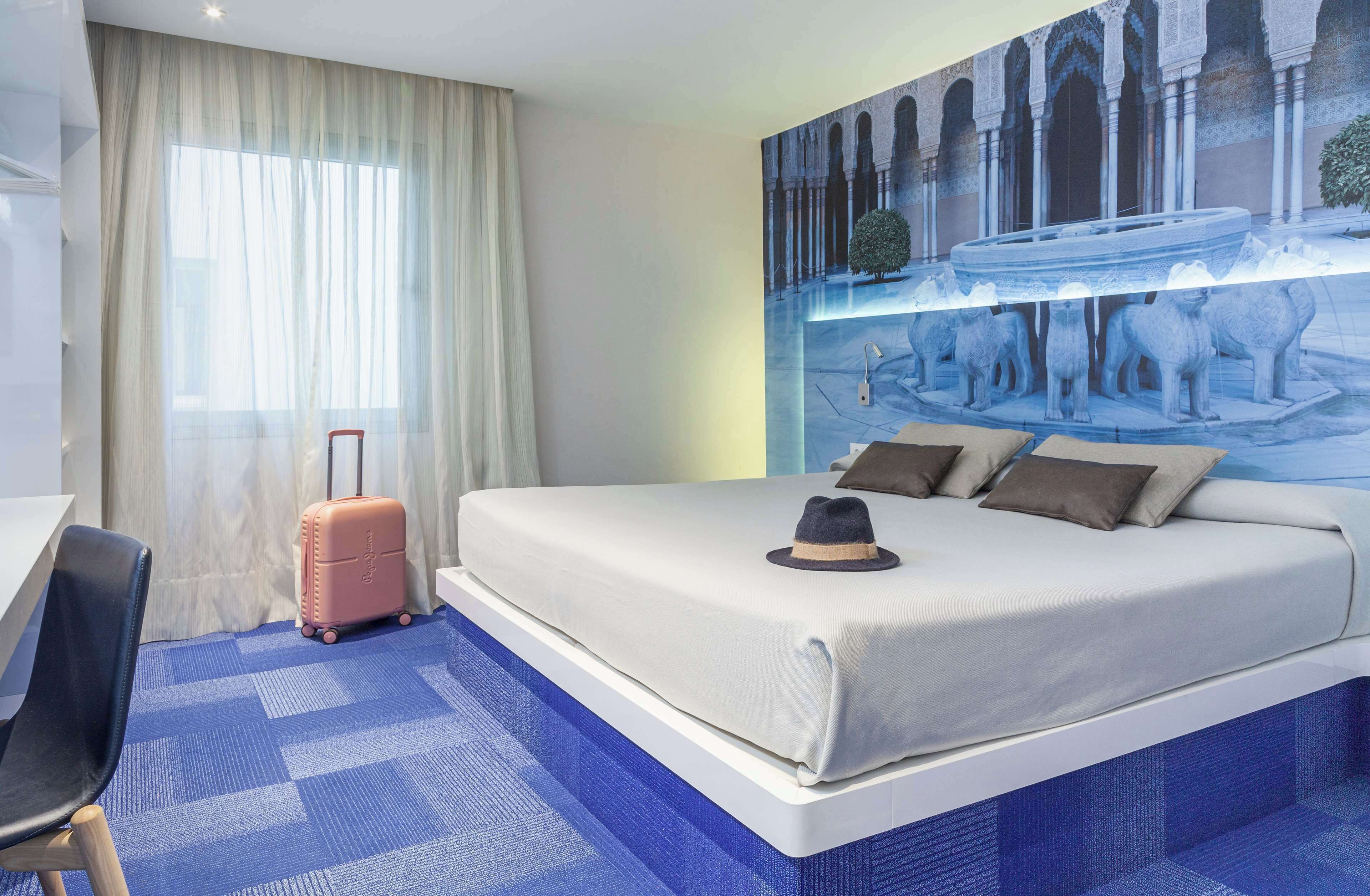Une chambre d’hôtel avec un grand lit, un bureau, une chaise, une valise et une fresque murale.