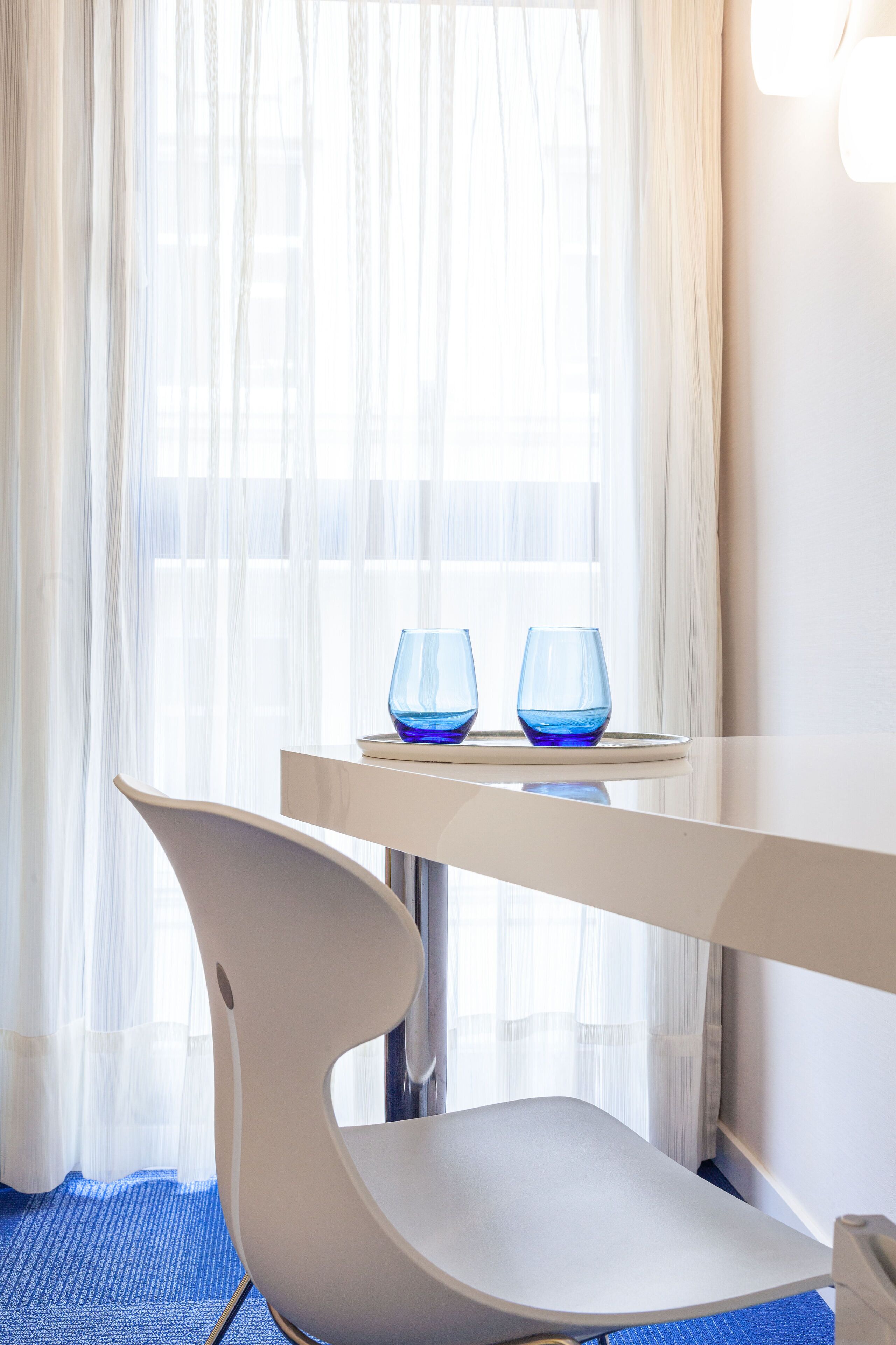 A modern room with a white table, two blue glasses, a white chair, and sheer curtains.