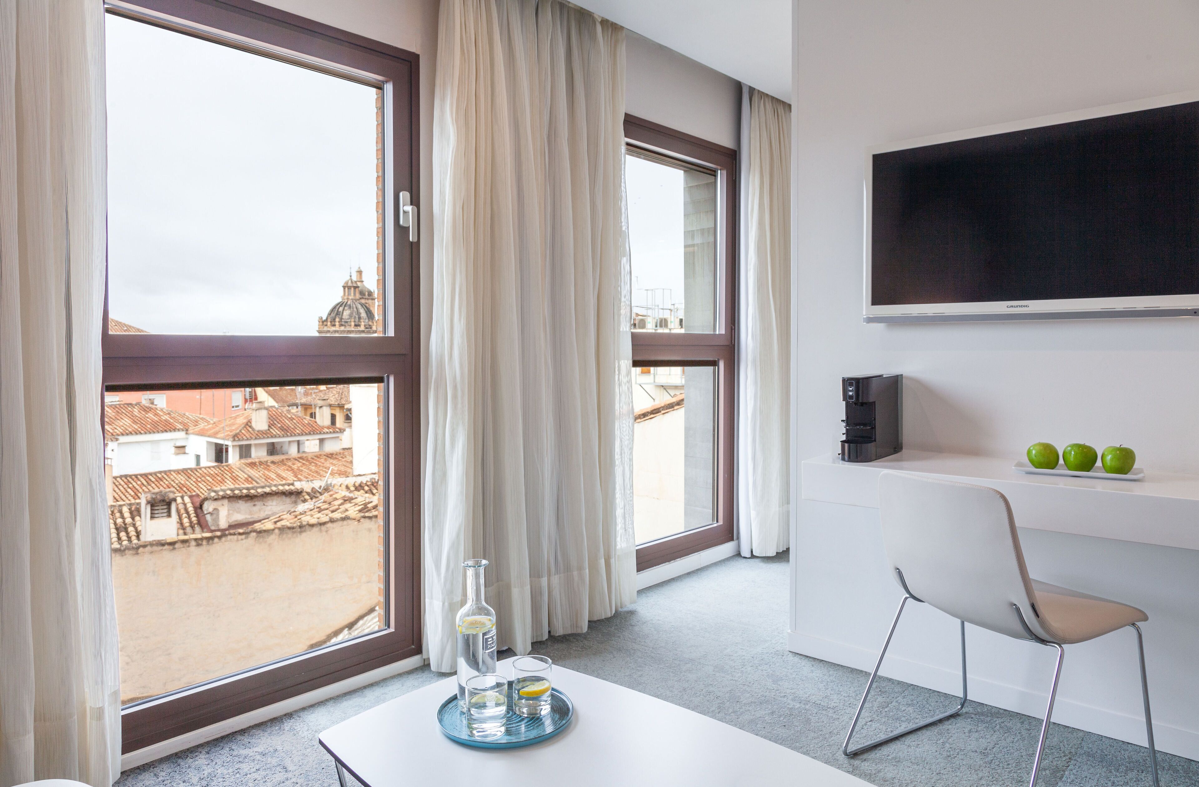 A hotel room with large windows, a flat-screen TV, a table with a bottle and glasses, and a chair.