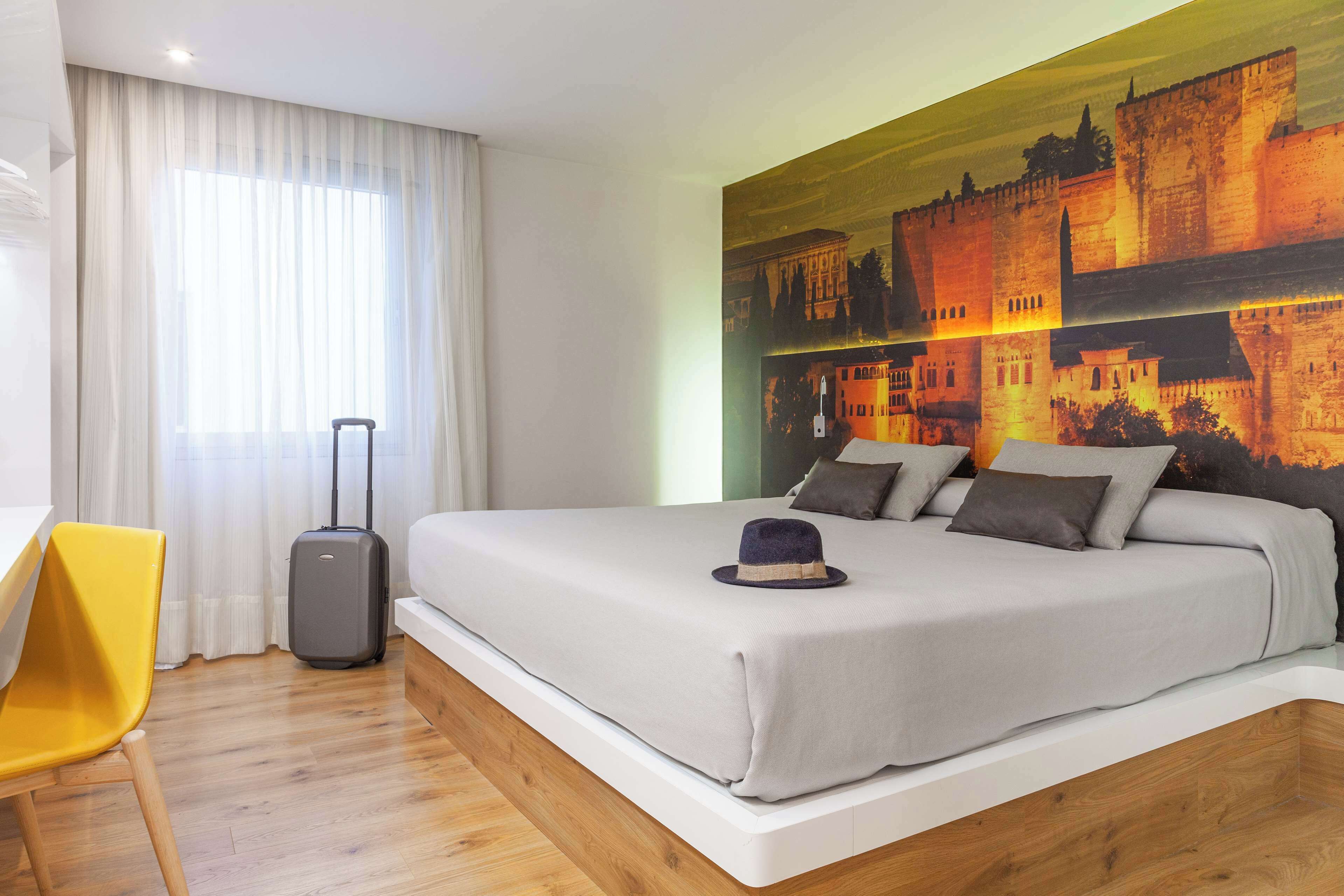 A hotel room with a large bed, a suitcase, a yellow chair, and a mural of a castle on the wall.