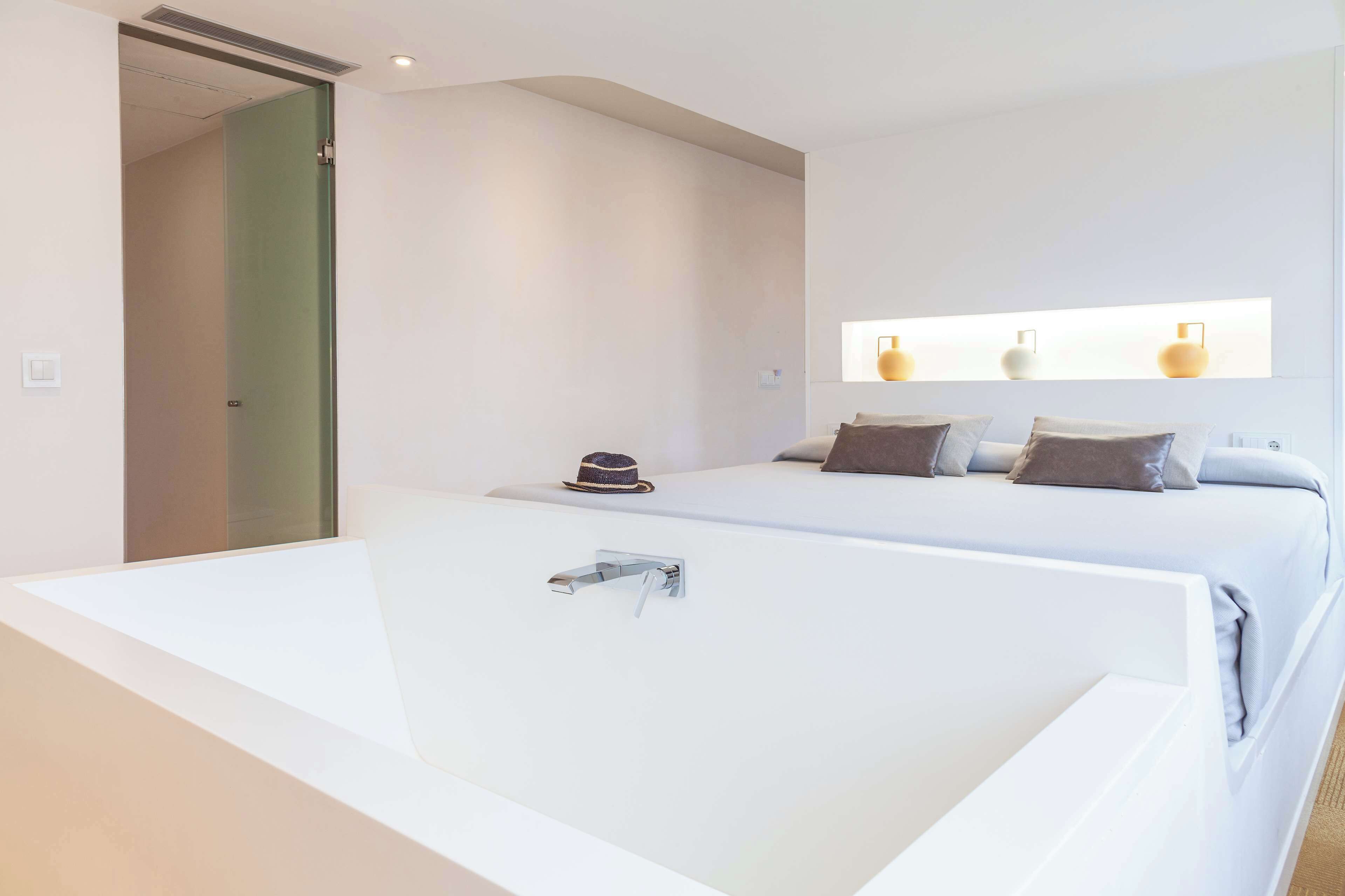 A modern hotel room with a large white bathtub, a bed with multiple pillows, and a hat on the bed.