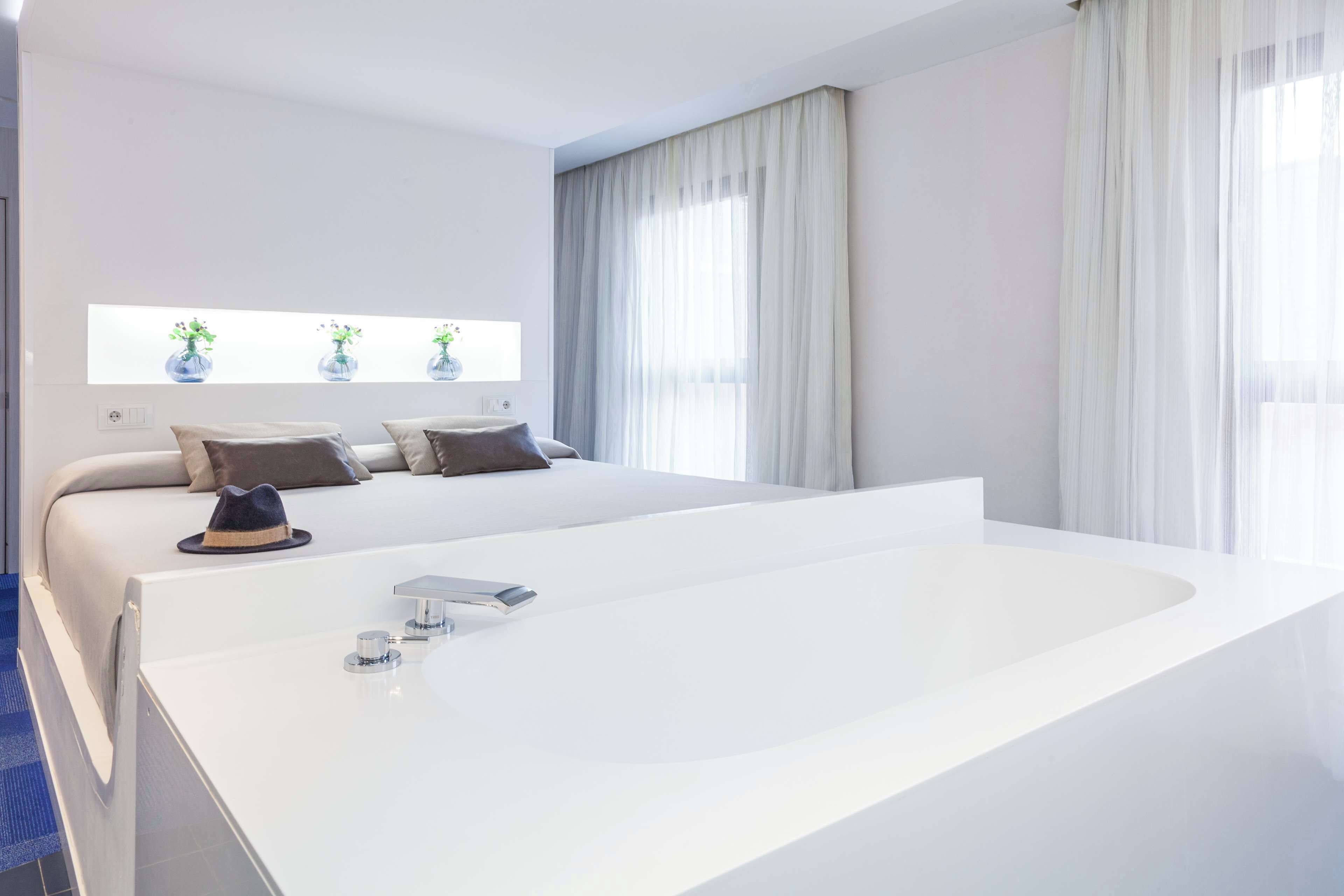 Ein modernes Schlafzimmer mit einer großen weißen Badewanne, einem Bett mit vielen Kissen und einem Fenster mit durchsichtigen Vorhängen.
