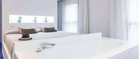 A modern bedroom with a large white bathtub, a bed with multiple pillows, and a window with sheer curtains.