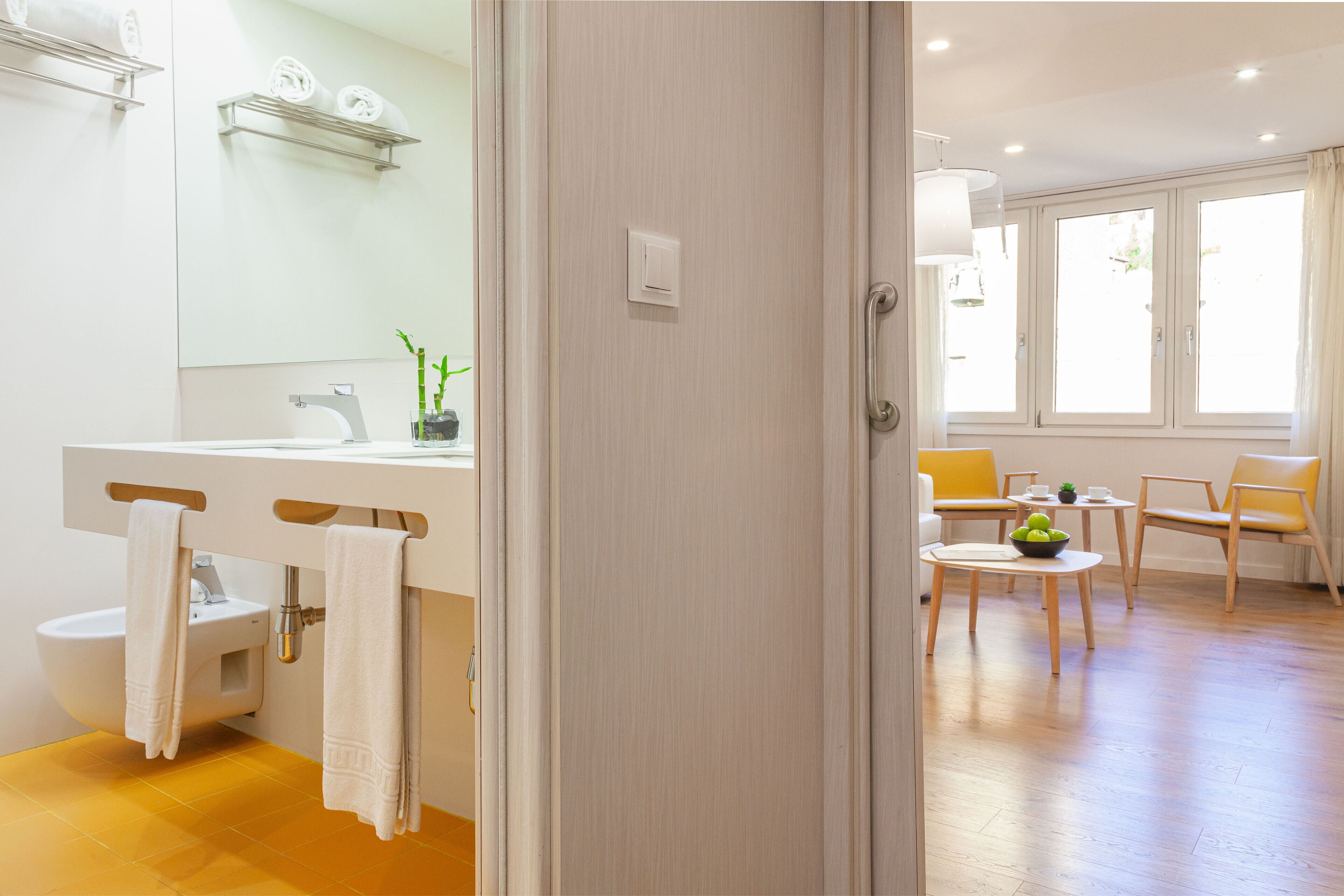 A modern bathroom with a sink, mirror, and towel rack. There is a dining area with a table and chairs, and a window with curtains.