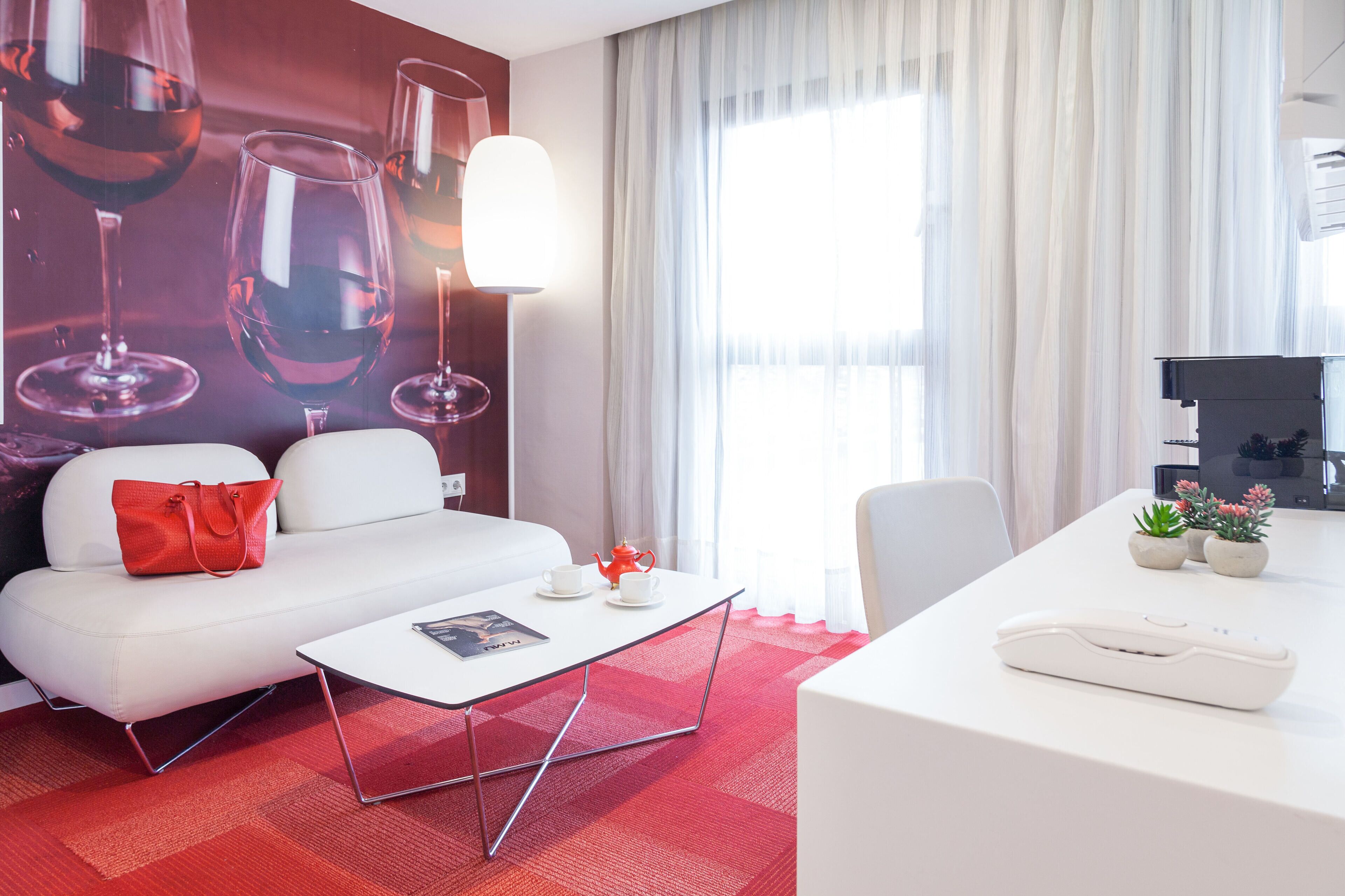 A modern hotel room with a white sofa, a small white table, a desk with a lamp, and a large window with sheer curtains.