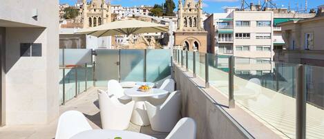 A rooftop terrace with white furniture, a table with fruit, and a view of historic buildings in the background.