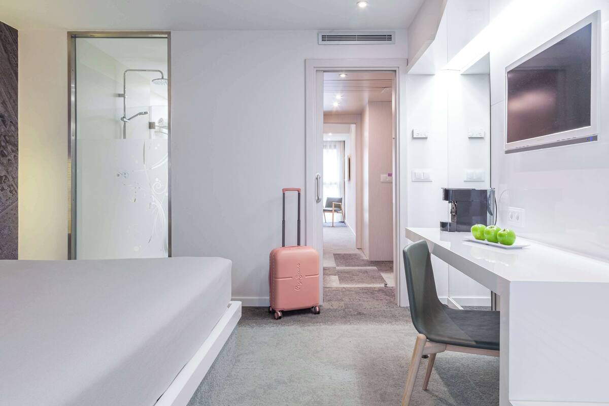 Ett modernt hotellrum med en säng, ett skrivbord, en stol, en resväska och en duschhörna.