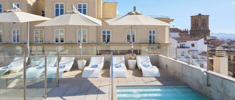A rooftop pool area with sun loungers and umbrellas, a clear view of a historic tower in the background, and a pool with a glass barrier.